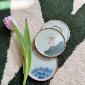 Può includere: Tre sottobicchieri in ceramica con sfondo blu chiaro e motivi floreali. I sottobicchieri presentano un fiore di loto con petali rosa e foglie verdi, e un fiore blu con accenti bianchi.