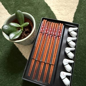 Pode incluir: Um conjunto de dez pares de pauzinhos de madeira com desenhos florais vermelhos e brancos, embalados em uma caixa preta com cinco apoios de pauzinhos em forma de coelho branco.