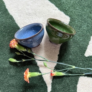 May include: Two small, ceramic bowls, one blue and one green, sit on a patterned rug. The bowls have a dark brown rim. A sprig of orange and yellow carnations lies between the bowls. The rug has a green and white design.