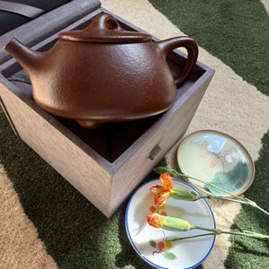 Könnte beinhalten: Eine braune Keramik-Teekanne mit Deckel, Henkel und Ausguss, in einer Holzkiste. Zwei kleine dekorative Teller und orangefarbene Blumen sind in der Nähe platziert. Die Teekanne hat eine satte, erdige Farbe.