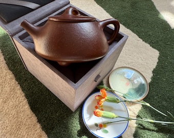 May include: A brown ceramic teapot with a lid, handle, and spout, resting inside a wooden box. Two small decorative plates and orange flowers are placed nearby. The teapot is a rich, earthy color.