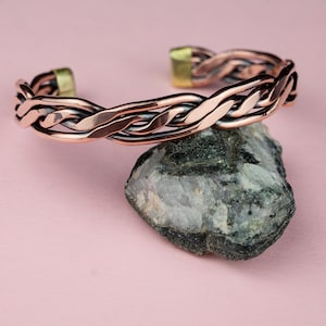May include: A copper-colored cuff bracelet with a braided design. The bracelet is resting on a gray rock against a pink background. The ends of the bracelet have gold-colored accents.