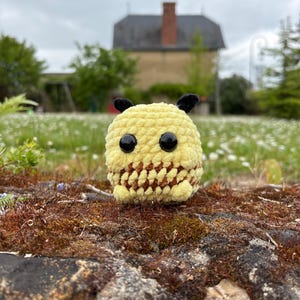 Puede incluir: Un juguete de peluche de abeja tejido a crochet con ojos negros y antenas, sentado en una roca cubierta de musgo. La abeja es amarilla y marrón con una textura peluda.