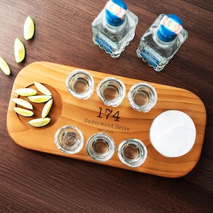 Puede incluir: Una bandeja de madera con seis vasos de chupito, un círculo de sal y una sección de gajos de lima. Dos botellas de tequila con etiquetas y tapones azules y blancos están en el fondo. La bandeja tiene el texto "174 Underwood Drive EST. 2025".
