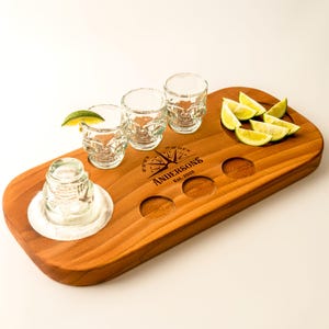 Puede incluir: Bandeja de madera con tres vasos de chupito, un soporte y gajos de lima. Los vasos de chupito tienen un diseño de calavera. La bandeja tiene el texto "Andersons Est. 2026" y un diseño de brújula.