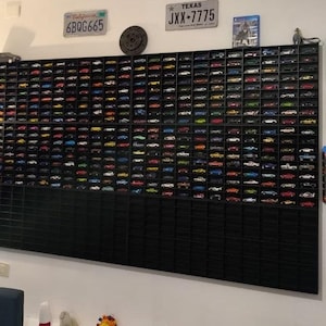 Könnte beinhalten: Eine große schwarze Vitrine mit zahlreichen Miniatur-Spielzeugautos in verschiedenen Farben. Die Vitrine ist an einer weißen Wand montiert, mit Nummernschildern und gerahmten Autokunstwerken in der Nähe. Eine Sammlung von Spielzeugautos.