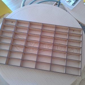 May include: A wooden display shelf with multiple small rectangular compartments. The shelf is made of light brown wood with darker brown edges. The compartments are arranged in a grid pattern, suitable for organizing small items.