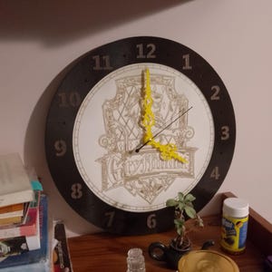 May include: A round wall clock with a black frame and a white face. The clock features the word "Gryffindor" and a crest design in the center. The clock hands are yellow. The numbers are black.