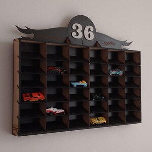 Puede incluir: Una estantería de pared de madera negra con treinta y seis compartimentos rectangulares individuales para coches de juguete. La parte superior presenta un cabecero decorativo negro con el número 36 en blanco y el logo de Hot Wheels.