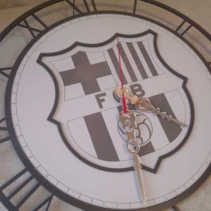 Puede incluir: Un reloj de pared redondo con el escudo del FC Barcelona en el centro. La esfera es blanca con números romanos negros. Las manecillas son plateadas y el segundero es rojo.