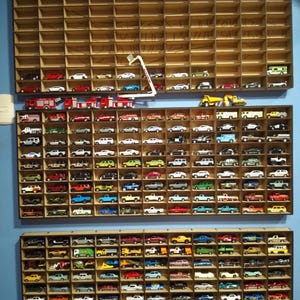 May include: A display of miniature toy cars in a wooden wall-mounted organiser with numerous compartments. The cars are in various colours and styles, including fire engines, saloons, and construction vehicles. A red double-decker bus sits on top.