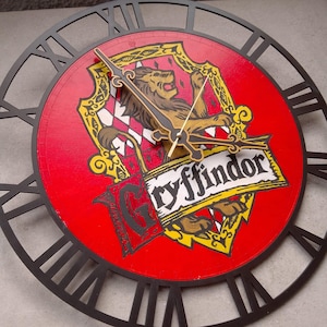 Può includere: Un orologio da parete circolare con sfondo rosso e un'illustrazione centrale dello stemma di Grifondoro. Lo stemma mostra un leone dorato, un motivo a scacchi rosso e bianco e la parola Gryffindor. Presenta numeri romani neri e lancette dorate sottili.