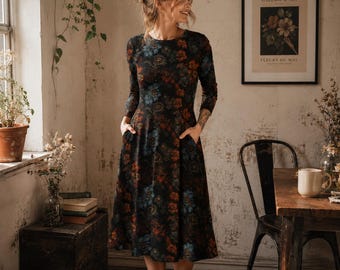 Puede incluir: Un vestido negro de manga larga con un estampado floral en tonos azules, naranjas y marrones. El vestido tiene cuello redondo y bolsillos. La mujer está de pie cerca de una ventana y una mesa con un cuadro enmarcado.