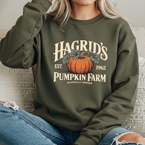 May include: An olive green sweatshirt featuring the text "Hagrid's Pumpkin Farm" with an illustration of an orange pumpkin and green leaves. The text "Est. 1962" and "Magically Grown" are also included in the design.