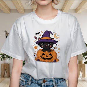 Op de afbeelding: Een wit T-shirt met korte mouwen en een grafische print van een zwarte kat met een paarse heksenhoed, zittend in een uitgesneden oranje pompoen. Het ontwerp bevat een sterrenstaf, een maansikkel, een vleermuis en herfstbladeren.