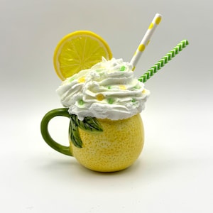 Puede incluir: Una taza de cerámica amarilla con forma de limón con hojas verdes y una cobertura de crema batida blanca. La taza tiene una pajita rayada blanca y amarilla y una pajita rayada verde y blanca que sobresalen de la crema batida.