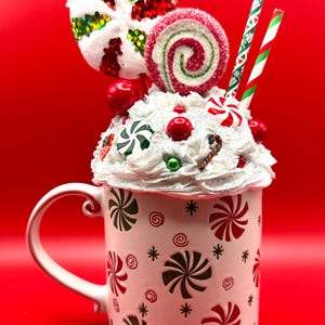 Puede incluir: Una taza festiva con base blanca, adornada con diseños de remolinos de menta roja y verde. La taza está rematada con un diseño de crema batida, adornada con caramelos, piruletas y pajitas decorativas, todo sobre un fondo rojo vibrante.