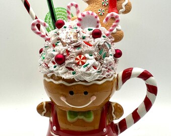 May include: A whimsical gingerbread man mug with a whipped cream topping, candy canes, and a gingerbread man cookie. The mug is brown with red accents and a smiling face. A festive holiday decoration.