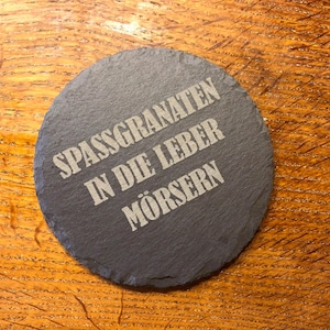 Puede incluir: Un posavasos redondo de pizarra gris oscuro con el texto alemán "SPASSGRANATEN IN DIE LEBER MÖSERN" grabado en blanco. El posavasos está sobre una superficie de madera con vetas naturales.