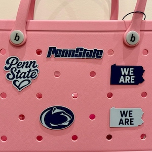 May include: A pink tote bag with multiple Penn State University themed patches. The patches include the words "Penn State" in a script font, the Penn State logo, and the words "WE ARE" in a state-shaped design.