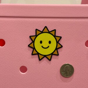 Puede incluir: Un bolso tote rosa con un dije en forma de sol amarillo. El dije tiene una cara sonriente y un contorno negro. Una moneda est&aacute; al lado del dije para comparar el tama&ntilde;o. La bolsa tiene varios agujeros redondos.