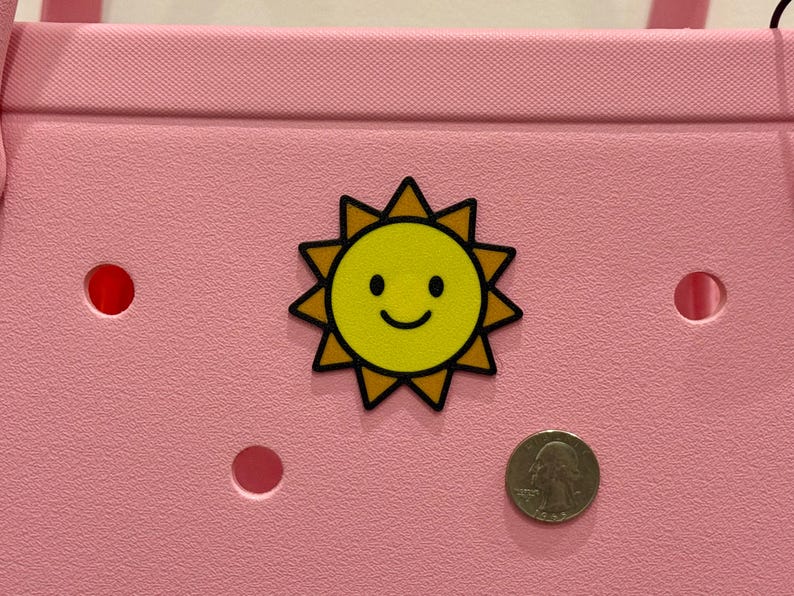 Puede incluir: Un bolso tote rosa con un dije en forma de sol amarillo. El dije tiene una cara sonriente y un contorno negro. Una moneda est&aacute; al lado del dije para comparar el tama&ntilde;o. La bolsa tiene varios agujeros redondos.