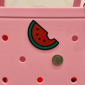 Puede incluir: Un bolso tote rosa con un dije de rebanada de sand&iacute;a. El dije es rojo con semillas negras y una c&aacute;scara verde. Una moneda est&aacute; al lado del dije. La bolsa tiene varios agujeros circulares rojos.
