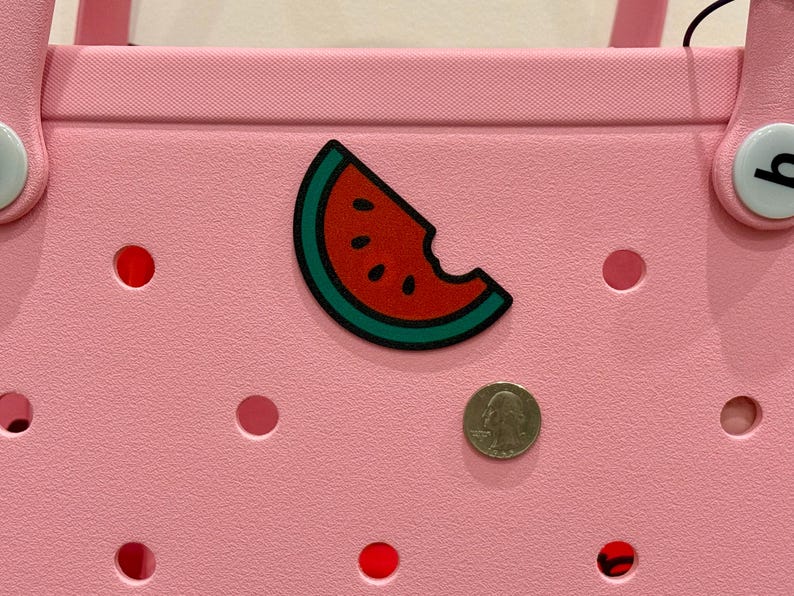 Puede incluir: Un bolso tote rosa con un dije de rebanada de sand&iacute;a. El dije es rojo con semillas negras y una c&aacute;scara verde. Una moneda est&aacute; al lado del dije. La bolsa tiene varios agujeros circulares rojos.