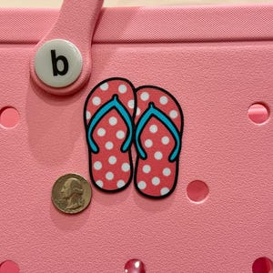 May include: Pink and white polka dot flip flops with blue straps. The flip flops are on a pink background with a white letter "b" on a round button.