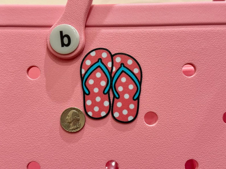 May include: Pink and white polka dot flip flops with blue straps. The flip flops are on a pink background with a white letter "b" on a round button.