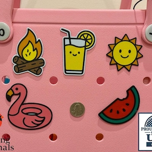 May include: Pink tote bag with summer-themed stickers, including a campfire, a juice with a lemon slice, a smiling sun, a flamingo float, a watermelon slice, and a quarter. The bag also has a "Proudly Made in the USA" sticker.