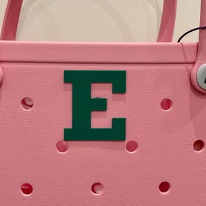 May include: Pink tote bag with a green letter "E" on the front. The bag has two pink handles and a series of circular cutouts. The handles are attached with white and black circular fasteners.
