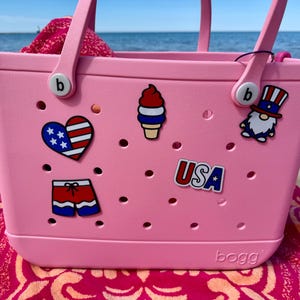 May include: A pink tote bag with various patriotic-themed charms, including a heart with the American flag, an ice cream cone, a gnome, and the word "USA". The bag is on a beach towel.