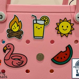 May include: A pink tote bag adorned with summer-themed charms, including a campfire, a juice drink, a smiling sun, a flamingo float, and a watermelon slice. A quarter coin is also present. The bag has a 'Proudly Made in the USA' sticker.