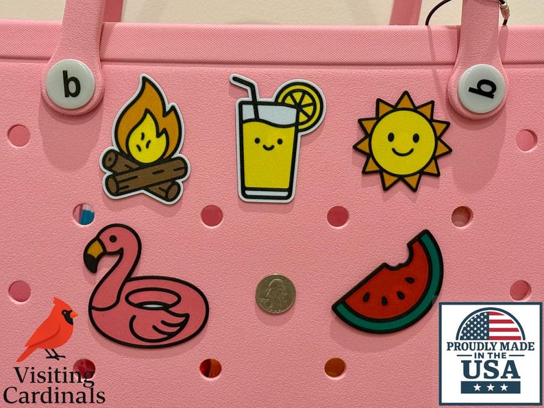 Puede incluir: Un bolso tote rosa adornado con amuletos con temas de verano, que incluyen una fogata, una bebida de jugo, un sol sonriente, un flotador de flamenco y una rebanada de sand&iacute;a. Tambi&eacute;n hay una moneda. La bolsa tiene una pegatina 'Hecho con orgullo en los EE. UU.'