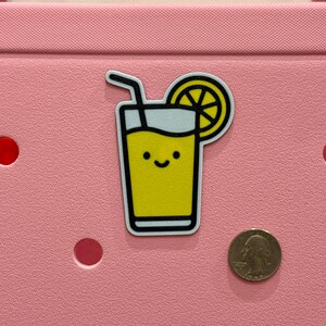 Puede incluir: Una pegatina de limonada amarilla con una cara sonriente, un contorno negro y una pajita. Una rodaja de lim&oacute;n est&aacute; en la parte superior derecha. La pegatina est&aacute; sobre una superficie rosa junto a una moneda.
