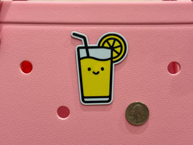 Puede incluir: Una pegatina de limonada amarilla con una cara sonriente, un contorno negro y una pajita. Una rodaja de lim&oacute;n est&aacute; en la parte superior derecha. La pegatina est&aacute; sobre una superficie rosa junto a una moneda.