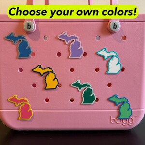 May include: A pink tote bag with colorful Michigan state-shaped charms. The charms are in various colors, including blue, purple, yellow, green, and pink. The text "Choose your own colors!" is displayed at the top.
