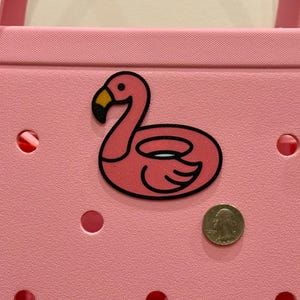 Puede incluir: Un bolso tote rosa con una pegatina de flotador con forma de flamenco. La pegatina tiene un contorno negro y un pico amarillo. Una moneda est&aacute; al lado de la pegatina para comparar el tama&ntilde;o. La bolsa tiene varios agujeros redondos.