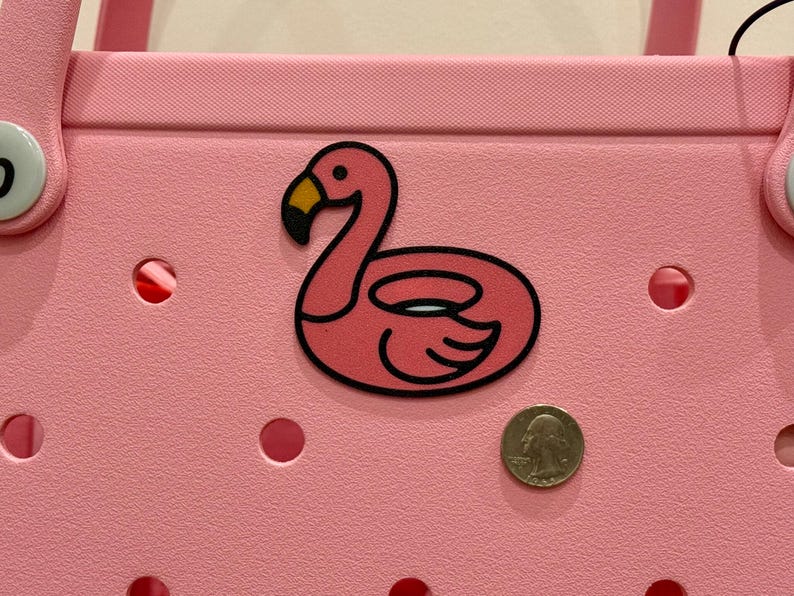 Puede incluir: Un bolso tote rosa con una pegatina de flotador con forma de flamenco. La pegatina tiene un contorno negro y un pico amarillo. Una moneda est&aacute; al lado de la pegatina para comparar el tama&ntilde;o. La bolsa tiene varios agujeros redondos.