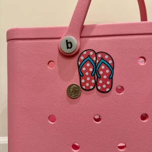 May include: Pink tote bag with a white handle and a white button with the letter "b" on it. The bag has a white and red polka dot flip flop sticker on it.