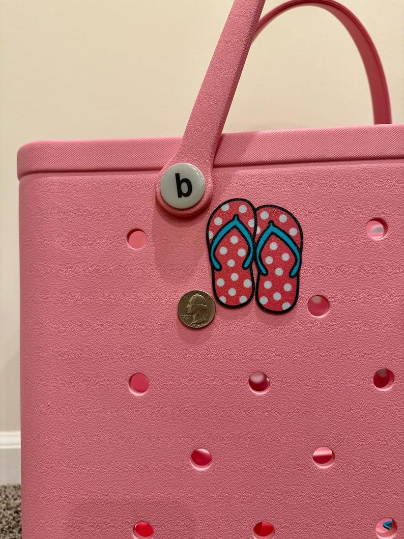 May include: Pink tote bag with a white handle and a white button with the letter "b" on it. The bag has a white and red polka dot flip flop sticker on it.