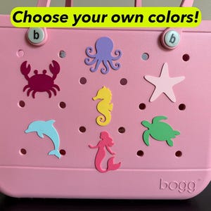 May include: Pink tote bag with various sea creature cutouts, including a crab, octopus, seahorse, starfish, dolphin, mermaid, and turtle. The bag has the text "Choose your own colors!" and the brand name "bogg".