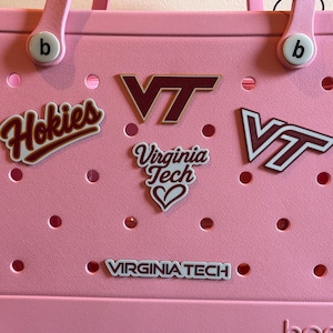 May include: A pink tote bag adorned with multiple Virginia Tech-themed decals. The decals include the word "Hokies", the letters "VT", and the words "Virginia Tech" with a heart symbol. The bag has a handle and a series of circular holes.