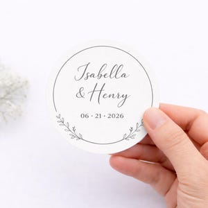 May include: A circular, off-white sticker featuring the names "Isabella & Henry" in cursive, and the date "06.21.2026". A subtle floral design adorns the bottom. The sticker is held by a hand.