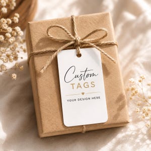 May include: A brown rectangular gift box tied with twine and a bow. A white tag with the words "Custom TAGS" in black and gold lettering is attached to the box. The tag also includes the text "YOUR DESIGN HERE".