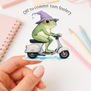 May include: A sticker featuring a green frog wearing a purple wizard hat, riding a white scooter. The text "Off to commit tom foolery" is above the frog. The frog has large red eyes and is smiling.