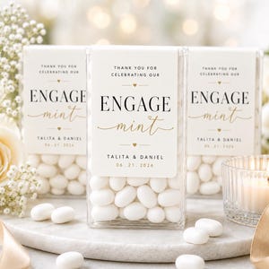 Peut inclure: Trois contenants rectangulaires transparents remplis de menthes ovales blanches. Chaque contenant porte une étiquette blanche avec du texte noir et doré indiquant "THANK YOU FOR CELEBRATING OUR ENGAGE mint TALITA & DANIEL 06.21.2026" avec des petits cœurs.
