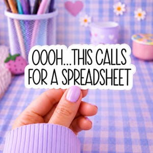 May include: A white die-cut vinyl sticker featuring a black border and the black text "OOOH...THIS CALLS FOR A SPREADSHEET" in a clear, sans-serif font. The sticker is held against a light purple background with a grid pattern and various desk items.