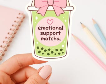May include: A die-cut sticker shaped like a light green matcha bubble tea cup. The cup features a pink bow at the top, a pink circular label with a heart, and the text "emotional support matcha." with dark green boba pearls at the base.